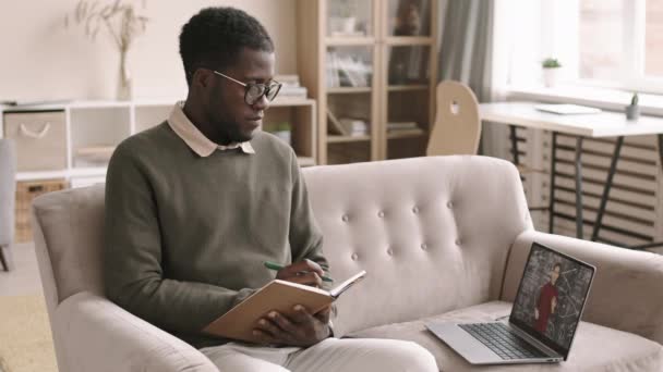 自宅のソファに座ってノートパッドでノートを作りながら ラップトップを使ったビデオ幾何学のレッスンを受けている若いアフリカ系アメリカ人の男性の中程度のショット — ストック動画