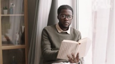 Orta boy bir Afrikalı genç adam günlük kıyafetler ve gözlük takıyor pencere kenarında oturuyor ve kitap okuyor.