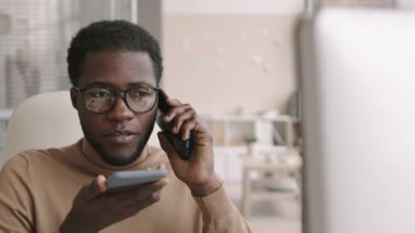 Orta boy Afro-Amerikan iş adamının şık günlük kıyafetler ve gözlükler giyip ofiste otururken ve aynı anda iki cep telefonu kullanırken çekilmiş bir fotoğrafı.
