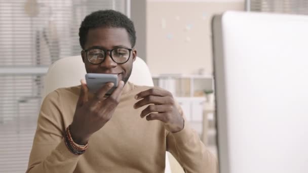 スマートフォンを手に職場に座り電話で話すカジュアルな服や眼鏡を身に着けている若いアフリカのビジネスマンの中程度のショット — ストック動画