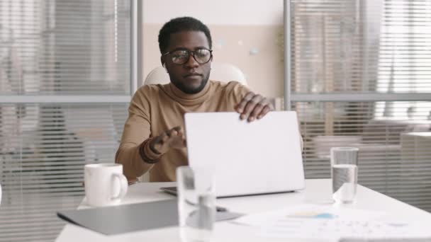 オフィスのデスクトップに座ってノートパソコンを閉じて目を閉じて座っている若いアフリカ系アメリカ人男性のオフィス労働者の中程度のショット — ストック動画
