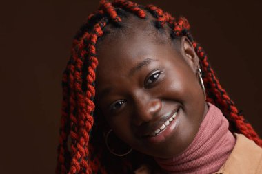 Saçları örgülü, koyu kahverengi arka planda poz verirken mutlu bir şekilde gülümseyen şık Afro-Amerikan kadın portresi.