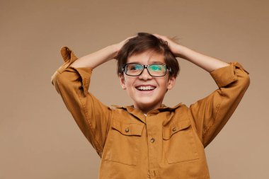 Stüdyoda bej arka planda dururken tasasız, gözlüklü ve mutlu bir şekilde gülümseyen çocuk portresi.