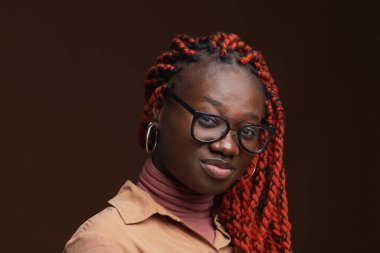 Saçları örülmüş, stüdyoda koyu kahverengi arka planda poz verirken kameraya bakan çağdaş Afro-Amerikan kadının yakın portresi.