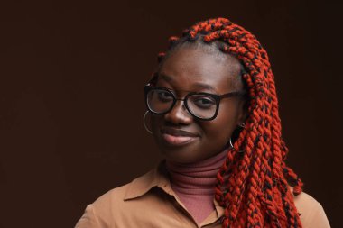 Saçı örgülü, kameraya gülümseyen ve stüdyoda koyu kahverengi arka planda poz veren kendine güvenen Afro-Amerikan kadının yakın portresi.