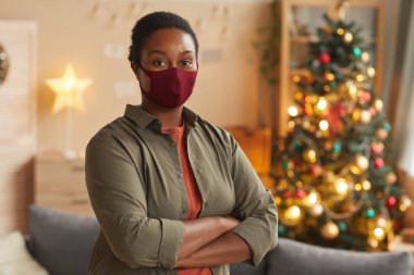 Waist up portrait of young African-American woman wearing mask and looking at camera while celebrating Christmas, copy space clipart