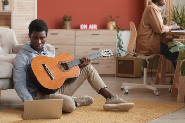 Gitar çalan ve dizüstü bilgisayar kullanan genç Afrikalı Amerikalı müzisyenlerin tam boy portresi.