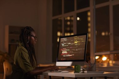 Side view portrait of contemporary African-American woman looking at computer screen and writing code while working late at night, copy space clipart
