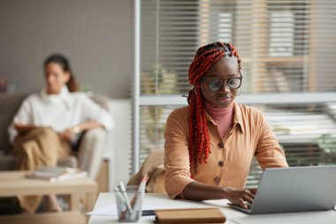 Ofiste arka planda insanlarla otururken dizüstü bilgisayar kullanan genç Afro-Amerikan kadın portresi, boşluğu kopyala