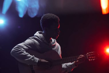 Sahnede renkli ışıklarla akustik gitar çalan genç Afro-Amerikalı bir adamın yan çizgisi.