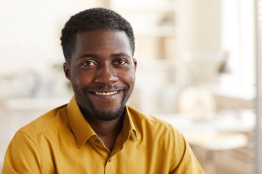 Beyaz ofis içinde çalışırken kameraya bakan gülümseyen Afro-Amerikalı adamın kafa ve omuz portresi.