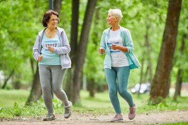Şehir parkında birbirleriyle konuşarak maraton koşan iki yaşlı kadının tam vücut fotoğrafı.