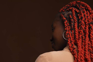 Saçları örgülü, koyu kahverengi arka planda poz verirken gözlerini kaçıran şık Afro-Amerikan kadının arka plan portresi.