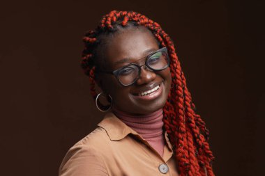 Örgü örmüş saçlarıyla kameraya gülümseyen kaygısız Afro-Amerikan kadın portresini yakından çek. Stüdyoda koyu kahverengi arka plana poz verirken, boşluğu kopyala.