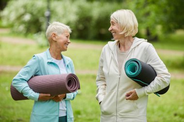 Spor kıyafetleri giymiş yoga minderleri tutan iki neşeli kadının orta boy portresi parkta birlikte vakit geçiriyorlar.