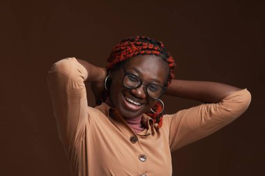 Saçları örgülü, kameraya bakarken mutlu bir şekilde gülümseyen ve stüdyoda koyu kahverengi arka planda poz veren kaygısız Afro-Amerikan kadın portresi.