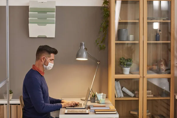 사무실에서 책상에서 일하는 마스크를 착용하고 노트북을 사용하는 남성의 측면시야 — 스톡 사진