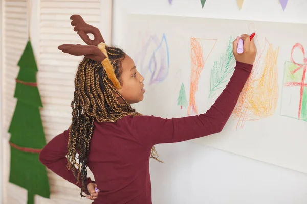 Waist up portrait of African-American girl wearing deer antlers and drawing on walls while enjoying art class on Christmas, copy space