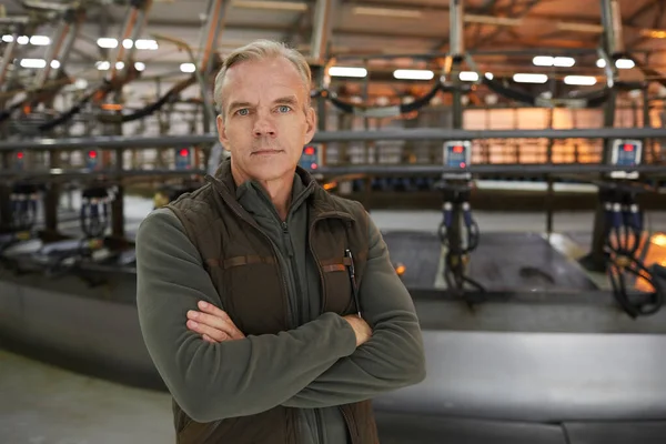 Waist up portrait of handsome mature man looking at camera while standing with arms crossed against industrial cow milking machine at modern dairy farm, copy space