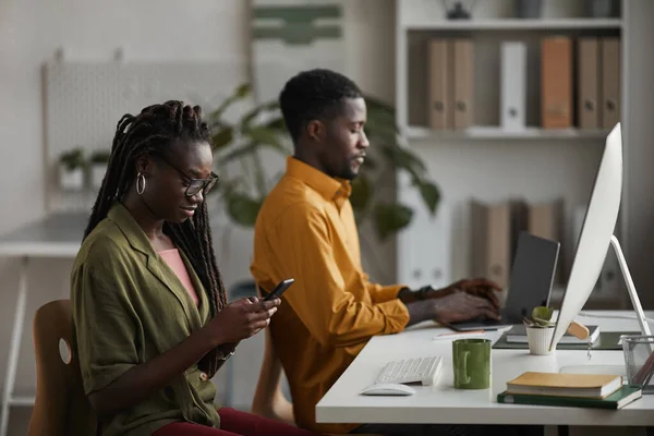 オフィスで働く2人の若者の肖像画 前面にスマートフォンを使用してエレガントなアフリカ系アメリカ人女性に焦点を当てています — ストック写真