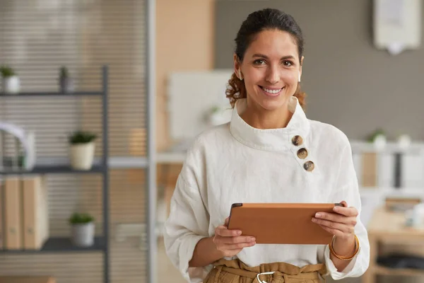 Waist up portrait of elegant modern businesswoman holding tablet and looking at camera with wireless earphones while standing in office, copy space