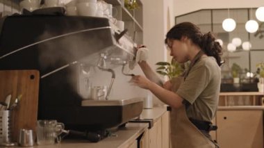 Geniş profesyonel espresso makinesinde buhar çubuğu kullanan beyaz kadın baristanın orta açıdan görünüşü nemli temizleme, arka planda yürüyen erkek iş arkadaşı.
