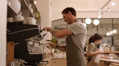 Kafkasyalı orta yaşlı bir adamın bardaki büyük bir profesyonel espresso makinesinde buhar çubuğu kullanırken, onu temizlerken, suyu silerken, kadın iş arkadaşının yanından geçerken ve giderken yan görüntüsü.