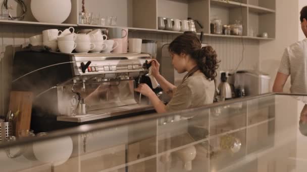 在咖啡店的大型专业咖啡机上 用蒸汽棒擦拭卷发的女性咖啡师的侧视图 然后将其清洁干净 将液体擦掉 中年白种人同事和她说话 面带微笑 — 图库视频影像