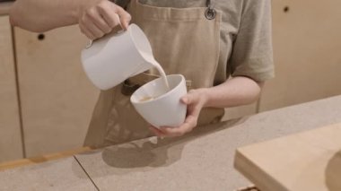 Tezgahta dikilen önlük giymiş, köpüklü sütü espresso ile bardağa dolduran ve latte üzerine şablon yapan tanınmamış baristanın yüksek açılı yakın çekimi.