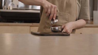 Tezgahın yanında duran tanınmayan baristanın yakın çekimleri iskele filtresindeki topraklanmış kahveye kurcalayıcı ile basıyor ve sonra geri dönüyor.