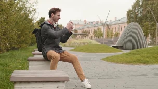 全景照片 身穿便衣的快乐白人男子 坐在公共广场的小长椅上 用看不见的东西的智能手机拍照 然后笑着看照片 — 图库视频影像