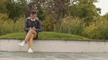 Kafkasyalı bir adamın rüzgarlı bir parktaki bankta otururken, etrafa bakınırken, beklerken, beklerken, akıllı telefon kullanırken çekilmiş uzun bir fotoğrafı.