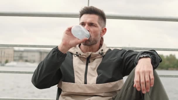 手すりにもたれかかる堤防に座ってスポーツ服を着ている中年の白人男性のパンニング スポーツボトルから水を飲んで カメラを見て — ストック動画