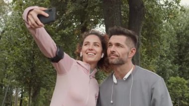 Orta yaşlı Kafkas sporcusu ve spor giyimli kıvırcık saçlı sporcunun parkta koşarken cep telefonuyla selfie çekerken orta boy fotoğrafı.