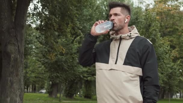 公園に立っている無線イヤフォンを身に着けている中年の白人スポーティな男性の中程度のショット 水の一口を作ります スマートウォッチで時間をチェックし 実行し続けます — ストック動画