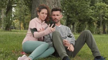 Orta yaşlı beyaz erkek ve spor giyinen kıvırcık saçlı atletik kadının parkta çimlerin üzerinde otururken ve cep telefonuyla selfie çekerken.
