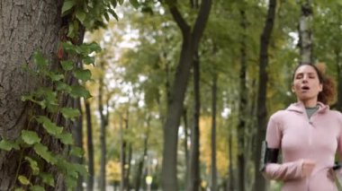 Orta boy beyaz kadın spor kıyafetler giymiş ormanda koşuyor, ağacın yanında duruyor, bir yudum su içiyor ve koşmaya devam ediyor.