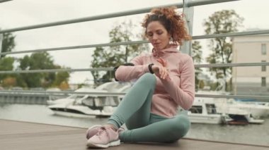 Spor kıyafetleri ve spor ayakkabıları giyen beyaz bir kadın iskelede oturuyor ve akıllı bakışlarını kontrol ediyor.