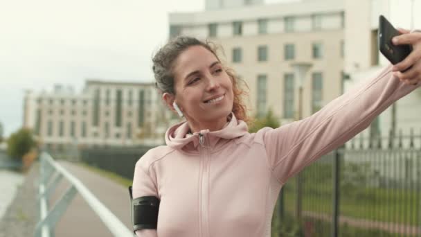 中镜头 漂亮的白人女赛跑选手穿着运动服和无线耳机 跑步后站在路基上 用电话自拍 — 图库视频影像