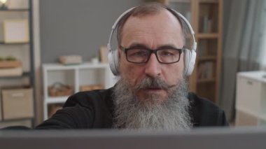 Ağır sakal, gözlük ve kulaklık takan, görünmez dizüstü bilgisayarın kapağını açan ve hayal kırıklığına uğrayan yaşlı beyaz adamın yakın çekimi.
