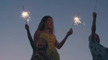 Uzun saçlı ve çocukları alacakaranlıkta okyanus kıyısında yan yana duran Bengal ışıklarını ellerinde tutan genç ve mutlu kadının alçak açılı görüntüsü.