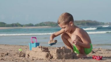 Sahilde oturan yeşil yüzme şortu giyen beyaz bir çocuk ve oyuncak kürek kemiğiyle kumdan kale yapıyor.