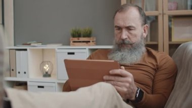 Orta boylu, beyaz, ağır sakallı ve bıyıklı bir erkek. Oturma odasında kanepeye yaslanmış, tablet bilgisayar kullanıyor, cep telefonuyla konuşuyor.