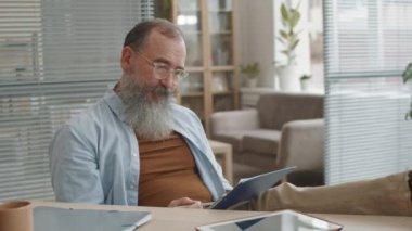 Orta yaşlı, uzun sakallı ve gözlüklü beyaz bir ofis çalışanı masanın üstünde oturmuş, panoda saçma sapan iş kağıtları okuyor, kafa sallıyor, çözüm düşünüyor.