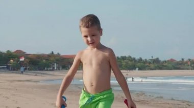 Küçük beyaz çocuk yeşil yüzme şortu giyiyor. Oyuncak kürek ve tırmıkla deniz kenarında kumdan kale yapıyor.