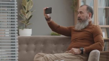 Orta uzun kenarlı, ağır gümüş sakallı, elinde akıllı bir telefonla video konferansında iletişim kuran yaşlı beyaz adamın görüntüsü.