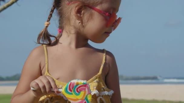 中等比例的白人小女孩 身穿黄色衣服 戴着心形太阳镜 在海滩上吃棒棒糖 — 图库视频影像
