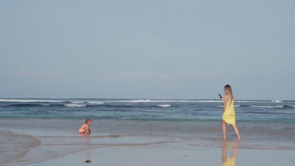 照片中年轻的妈妈穿着黄色的裙子 穿着长长的长发 沿着海滨散步 用电话为她可爱的小女儿拍照 — 图库视频影像