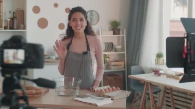 Beyaz kadın video blogcusu önlük takıyor, mutfakta masanın yanında duruyor, konuşuyor, kameranın önünde elini sallıyor ve gülümsüyor.