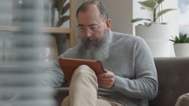 Orta boylu, gözlüklü, sakallı ve bıyıklı profesyonel bir beyaz erkek koltukta oturuyor, tablet kullanıyor, telefonla konuşuyor, gülümsüyor.